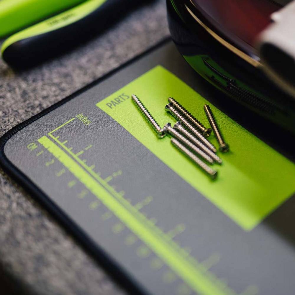 Close-up of a black and neon green parts measurement mat with several metal screws placed on it, photographed on a textured gray surface.