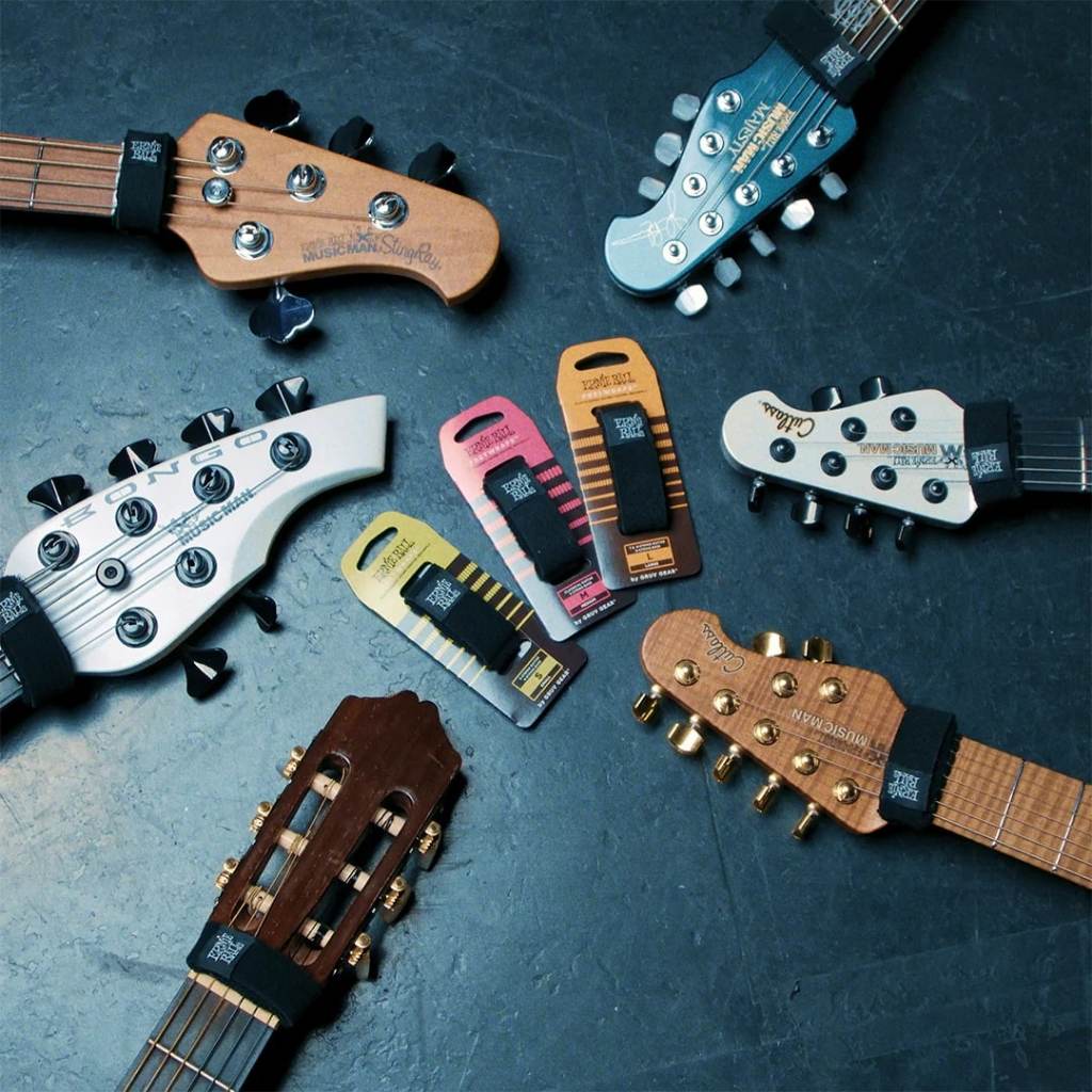 Six guitar foot pedals in assorted colors are arranged on a dark textured surface, with multiple guitar headstocks cropped around them to show the pedals in use. The pedals include black, pink, yellow, orange, and silver/black models, and the visible headstocks include brands such as Squier, Yamaha, and Ricken.
