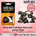 Ernie Ball heavy guitar picks in assorted black, white, and tortoiseshell colors, shown in their original retail packaging alongside a pile of loose picks on a pink background with product text and branding.
