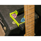 A close-up of a guitar pick set in a bright neon green tray, resting on a black guitar amplifier next to the neck of a wooden guitar. The set includes pink, blue, and yellow picks with black and white graphics, along with a black and yellow marker.